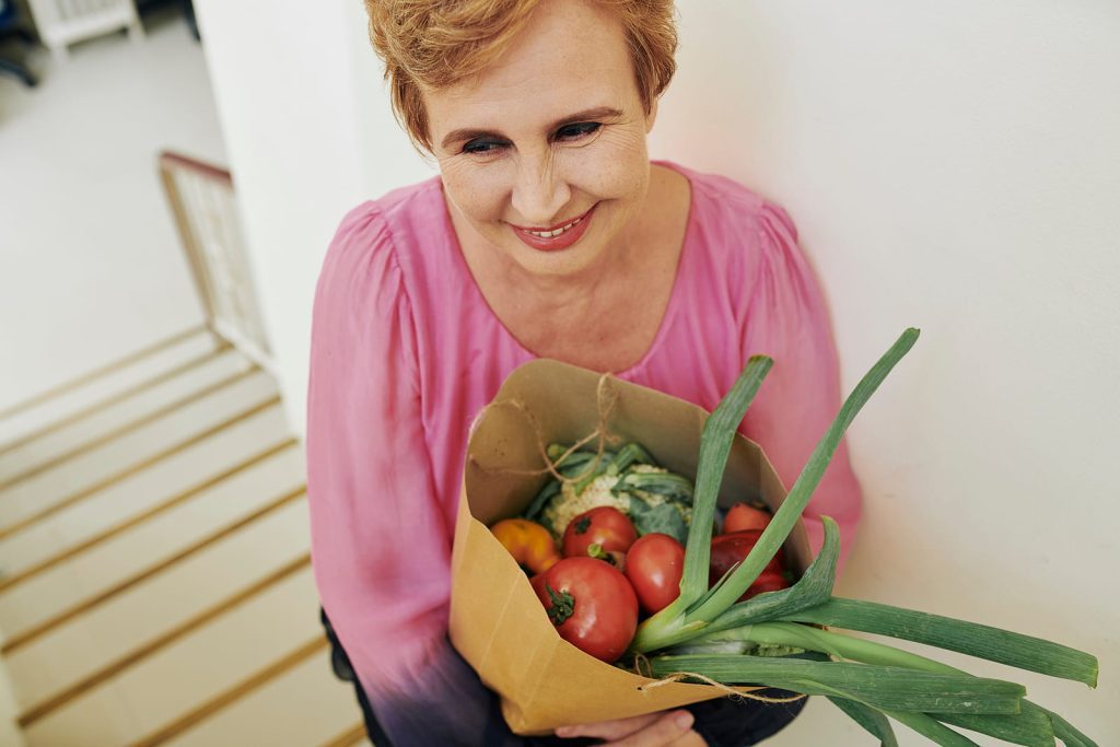 Woman in a pink blouse holding a paper bag filled with fresh vegetables, including tomatoes, peppers, and green onions, while standing indoors near stairs.